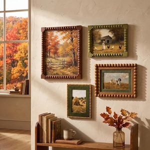 May include: Four rectangular wall frames with beaded edges displaying landscape and animal art. The frames are in shades of brown and olive green. Below, a wooden shelf holds stacked books, a ceramic mug, and a glass vase with autumn leaves.
