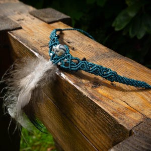 Peut inclure: Un collier en macramé turquoise avec un pendentif argenté et des perles noires, orné d'une plume grise et noire duveteuse. Le collier repose sur une surface en bois vieilli, mettant en valeur un style bohème.