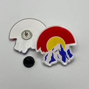 May include: Two Colorado state flag pins. One pin is white with a silver pin back. The other pin features a red, yellow, blue, and white design of the Colorado state flag. A black pin back is also included.