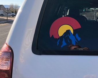 May include: A white SUV with a Colorado state flag sticker on the back window. The sticker features a red, yellow, and blue design with a mountain silhouette.