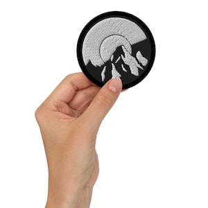 Pode incluir: Um remendo bordado preto e branco com uma cordilheira e uma lua cheia no fundo.