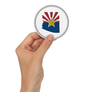 Könnte beinhalten: Ein weißer gestickter Aufnäher mit der Flagge des Bundesstaates Arizona. Die Flagge zeigt eine rote Sonne mit 13 Strahlen, einen blauen Hintergrund und einen gelben Stern in der Mitte.