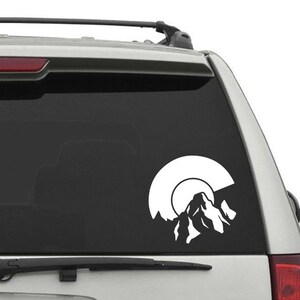 May include: White vinyl decal of a mountain range with a sun setting behind it. The decal is on the back window of a silver SUV.
