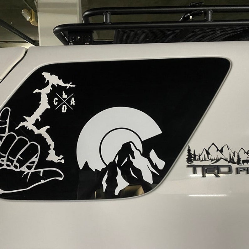 May include: A white car with black window decals. The decals feature a mountain range, a sun, a hand gesture, and a lake with the letters "CAD" inside a cross. The car also has a black "TRD PRO" emblem on the back.