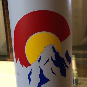 May include: A white water bottle with a sticker of the Colorado state flag. The sticker features a red circle with a yellow sun and a blue mountain range.