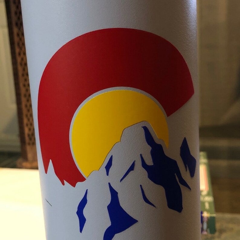 May include: A white water bottle with a sticker of the Colorado state flag. The sticker features a red circle with a yellow sun and a blue mountain range.