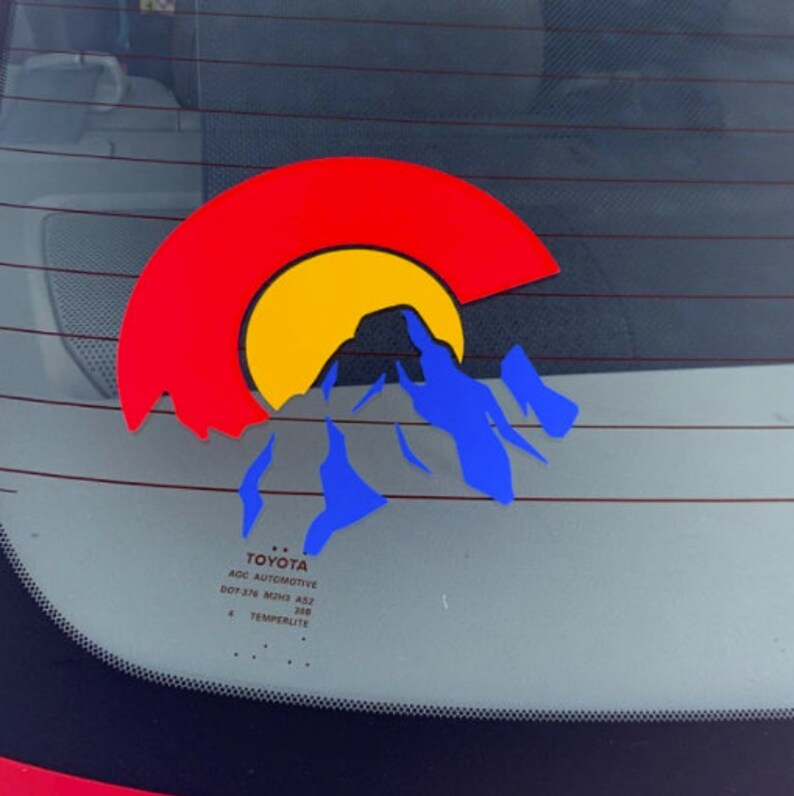 May include: A red, yellow, and blue Colorado state sticker on a car window. The sticker features a stylized mountain range with a sun setting behind it. The text "TOYOTA AGC AUTOMOTIVE DOT-3/8 M20x1.5 AS2 348 TEMPERLITE" is visible below the sticker.