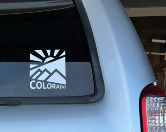 May include: A white sticker on the back window of a silver car. The sticker features a stylized sun rising over mountains with the word "COLORADO" below.