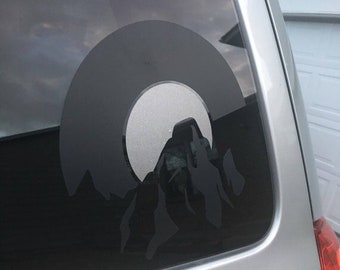 May include: A black and white silhouette of a mountain range with a circle around it, on a car window.