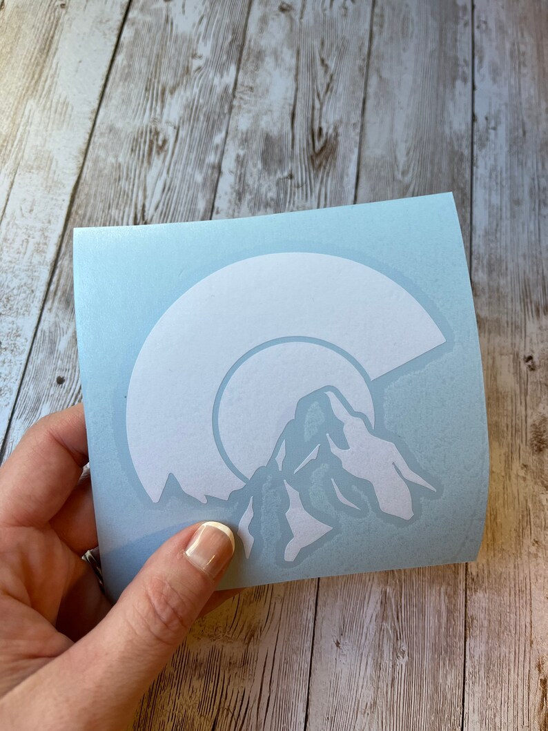 May include: White vinyl sticker with a mountain range silhouette and a crescent moon design. The sticker is on a light blue background.