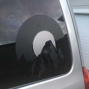 May include: A black and white silhouette of a mountain range with a circle around it, on a car window.