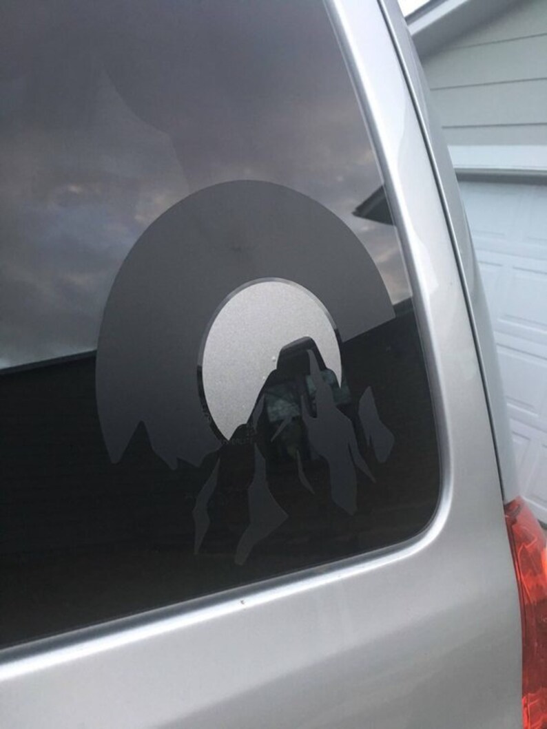 May include: A black and white silhouette of a mountain range with a circle around it, on a car window.