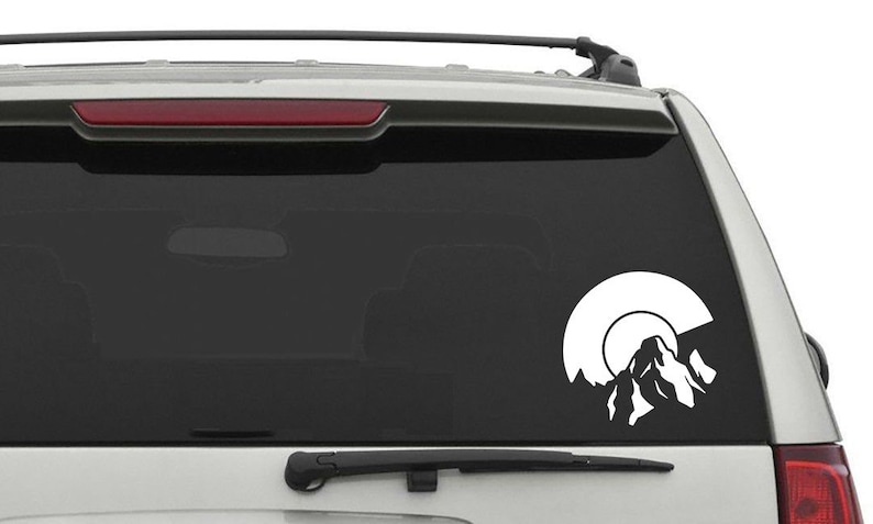 May include: White vinyl decal of a mountain range with a sun setting behind it. The decal is on the back window of a silver SUV.