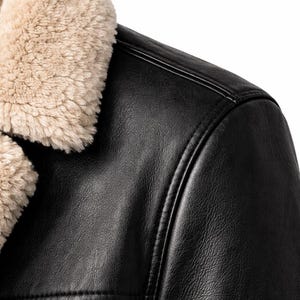 May include: Close-up of a black leather jacket with a beige faux fur collar. The jacket has a textured surface and visible stitching details along the seams and shoulders. The focus is on the upper portion of the jacket.