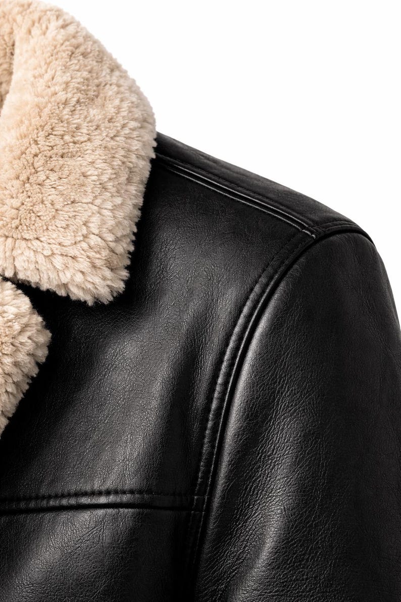 May include: Close-up of a black leather jacket with a beige faux fur collar. The jacket has a textured surface and visible stitching details along the seams and shoulders. The focus is on the upper portion of the jacket.