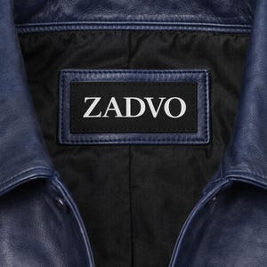 May include: Close-up of a navy blue leather jacket collar. The jacket has a black interior lining and a label with the white text "ZADVO". The leather has a textured appearance.