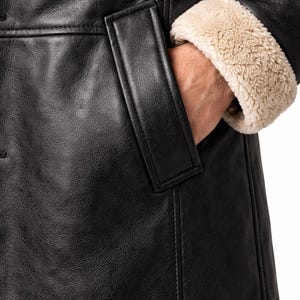May include: Close-up of a black leather coat with a pocket and a tan, fuzzy lining on the sleeve cuff. The coat has a classic design and appears to be made of high-quality materials. The focus is on the texture and details of the coat.