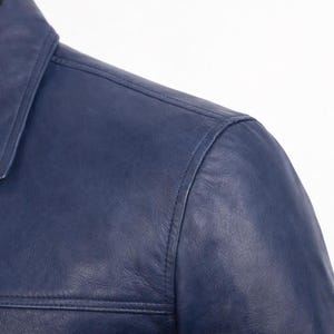 May include: Detail of a navy blue leather jacket. The image shows the collar and shoulder area, with visible stitching. The leather has a smooth finish and a subtle sheen. The jacket appears to be well-made.