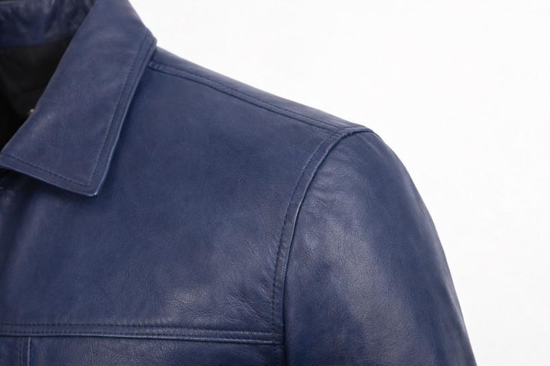 May include: Detail of a navy blue leather jacket. The image shows the collar and shoulder area, with visible stitching. The leather has a smooth finish and a subtle sheen. The jacket appears to be well-made.