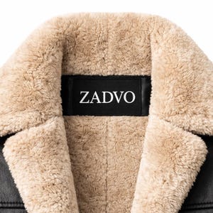 May include: Close-up of a black leather jacket with a beige shearling collar and lining. The brand name "ZADVO" is displayed on a black label on the collar.