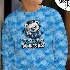Puede incluir: Una sudadera de manga larga con un patrón tie-dye azul que presenta un gráfico de un oso blanco con una sudadera con capucha azul y una gorra negra, señalando hacia adelante. Debajo del oso, el texto dice GANZ DÜNNES EIS en letras negritas.