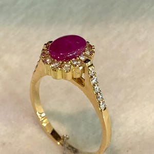 Może przedstawiać: Złoty pierścionek z dużym, owalnym rubinem otoczonym małymi, przezroczystymi kamieniami. Obręcz pierścionka jest również ozdobiona mniejszymi kamieniami. Rubin ma głęboki, czerwony kolor, a pierścionek ma polerowane wykończenie.