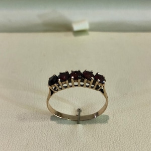 Op de afbeelding: Een gouden ring met vijf ovale, donkerrode edelstenen. De ring heeft een open ontwerp op de band. De edelstenen zijn gelijkmatig verdeeld over de bovenkant van de ring, tegen een neutrale achtergrond.
