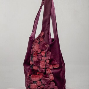 May include: A deep purple tote bag with a plush, textured design in shades of red and burgundy. The bag features long handles and a soft, velvety exterior. The decorative element is a cascade of textured, faux fur-like material.