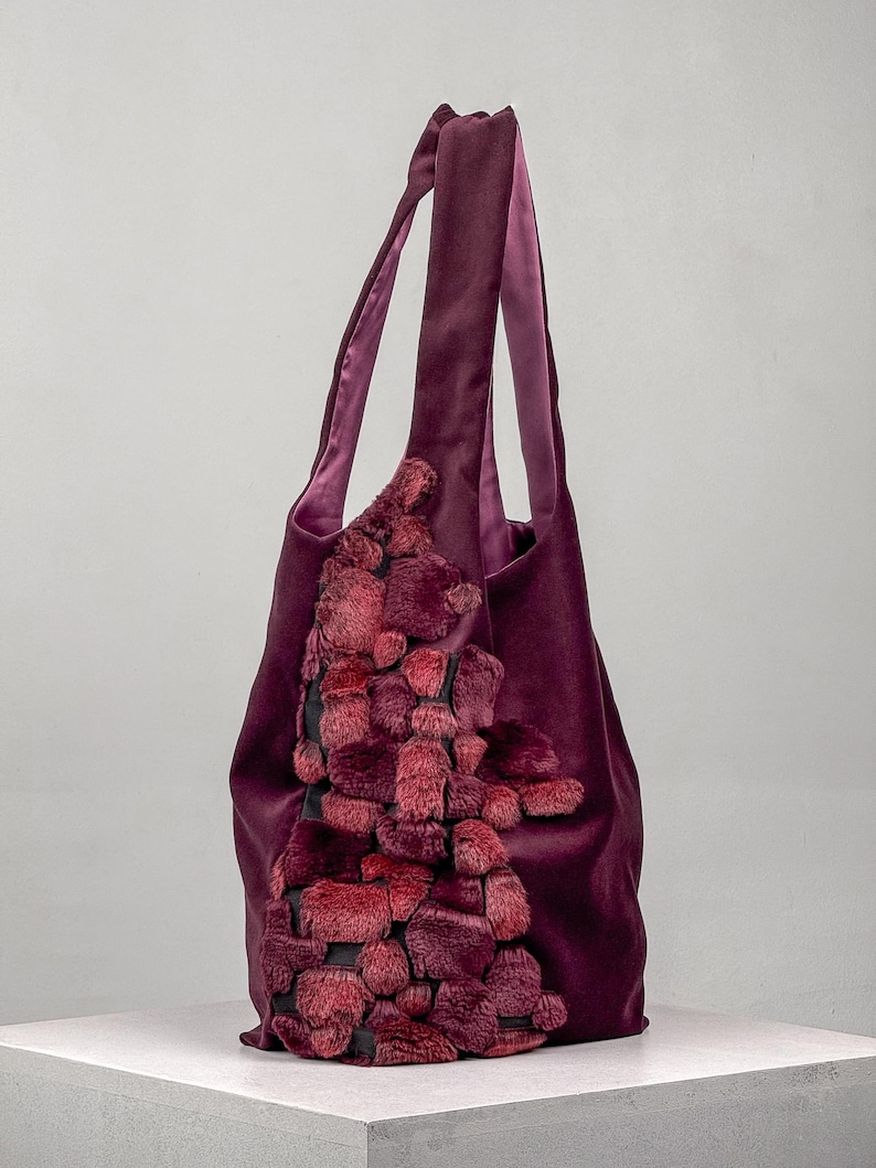 May include: A deep purple tote bag with a plush, textured design in shades of red and burgundy. The bag features long handles and a soft, velvety exterior. The decorative element is a cascade of textured, faux fur-like material.