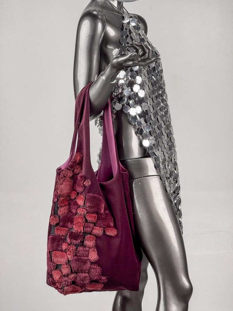 May include: A deep burgundy tote bag with a textured, raised pattern in shades of red and pink. The bag has a smooth, solid burgundy interior and handles. The bag is held by a mannequin wearing a silver sequin halter top.