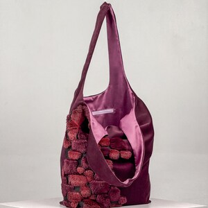 May include: A plum-colored tote bag with a plush, textured front panel in shades of red and burgundy. The bag has a soft, satin-like interior lining and long shoulder straps. The bag is sitting on a white surface.