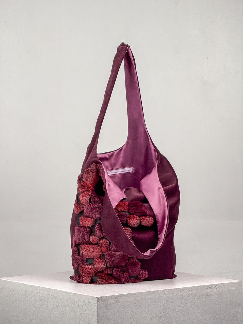 May include: A plum-colored tote bag with a plush, textured front panel in shades of red and burgundy. The bag has a soft, satin-like interior lining and long shoulder straps. The bag is sitting on a white surface.