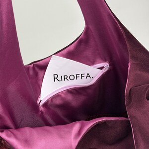 May include: A close-up of a deep purple tote bag with a matching interior. The bag features a zippered pocket with the text "RIROFFA." The bag's interior is a lighter shade of purple, and the exterior has a velvety texture.