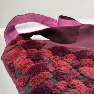 May include: A burgundy tote bag with a plush, textured design in shades of red. The bag features a satin handle and trim. The textured design is made of small, rectangular shapes. The bag is made of a soft material.