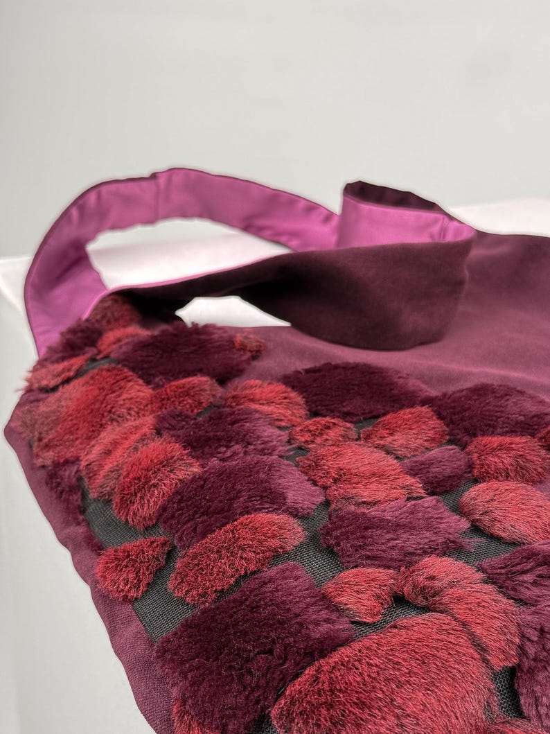 May include: A burgundy tote bag with a plush, textured design in shades of red. The bag features a satin handle and trim. The textured design is made of small, rectangular shapes. The bag is made of a soft material.