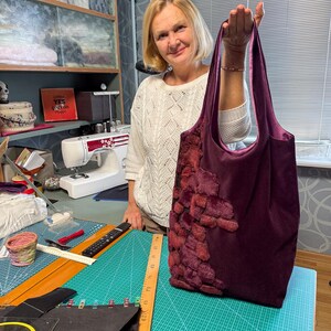 May include: A deep purple tote bag with a plush, textured design on the front. The bag has a soft, velvety appearance and a contrasting interior. The handles are a matching purple. The bag is held up by a person.