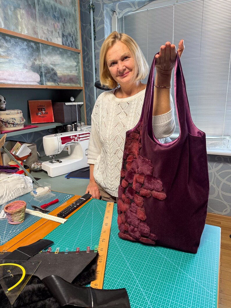 May include: A deep purple tote bag with a plush, textured design on the front. The bag has a soft, velvety appearance and a contrasting interior. The handles are a matching purple. The bag is held up by a person.