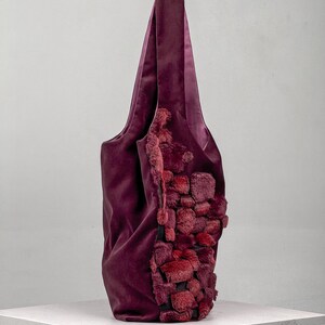 May include: A deep burgundy tote bag with a plush, textured design on one side. The bag features a soft, velvety material and a contrasting, textured accent in shades of red. The handles are long and made of the same burgundy material.