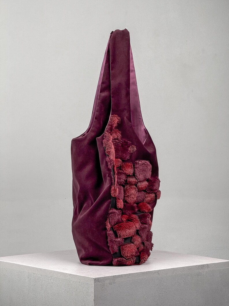 May include: A deep burgundy tote bag with a plush, textured design on one side. The bag features a soft, velvety material and a contrasting, textured accent in shades of red. The handles are long and made of the same burgundy material.