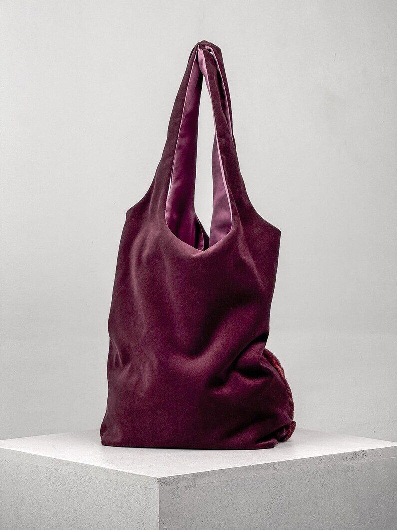 May include: A deep burgundy velvet tote bag with a contrasting pink satin lining. The bag has long handles and a soft, unstructured shape, perfect for carrying essentials. The bag is displayed on a white surface against a neutral background.