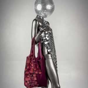 May include: A silver mannequin with a disco ball head and a sequined dress holds a burgundy tote bag. The bag features a textured, reddish-pink design. The mannequin is set against a gray background.