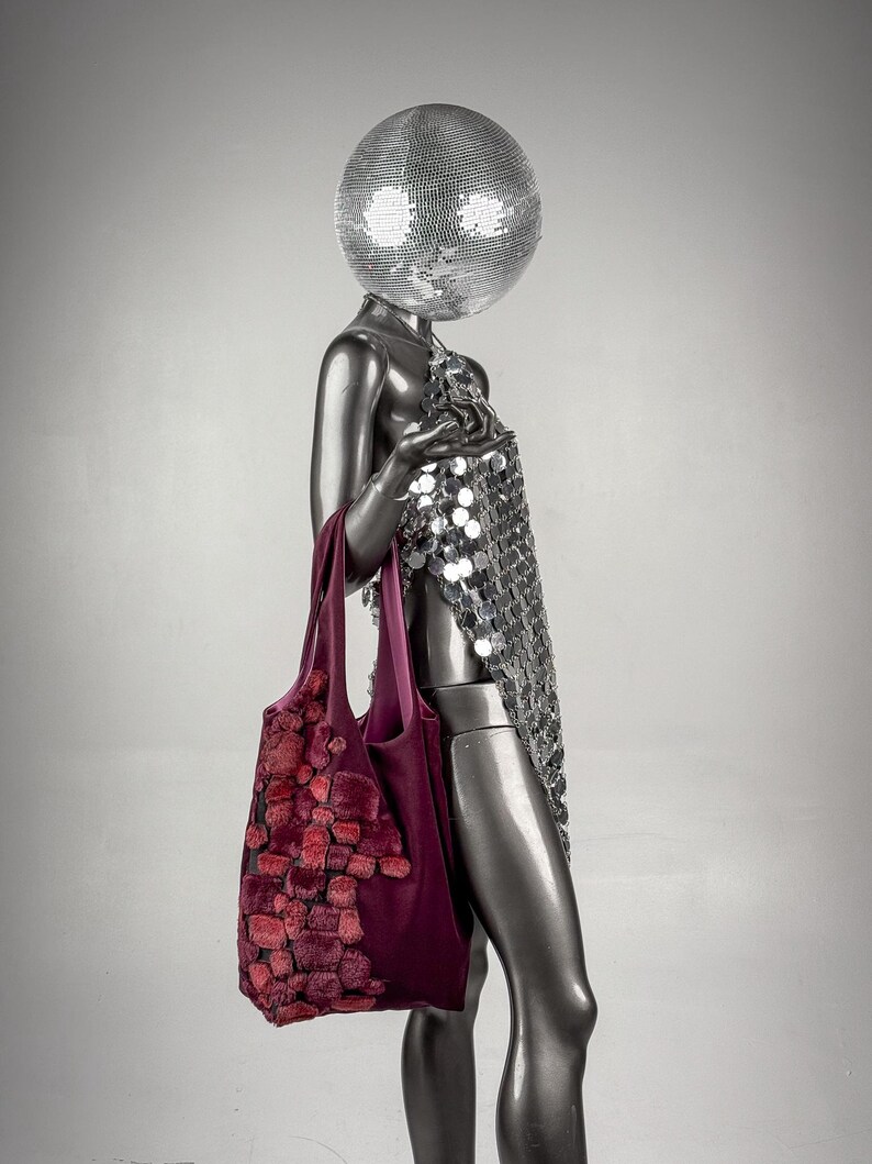 May include: A silver mannequin with a disco ball head and a sequined dress holds a burgundy tote bag. The bag features a textured, reddish-pink design. The mannequin is set against a gray background.