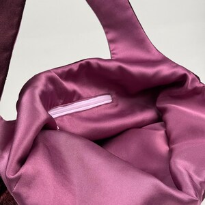 May include: A close-up of a tote bag with a deep burgundy exterior and a satin-like, mauve interior. The bag has a long strap and a zipper closure. The fabric has a soft, luxurious appearance.
