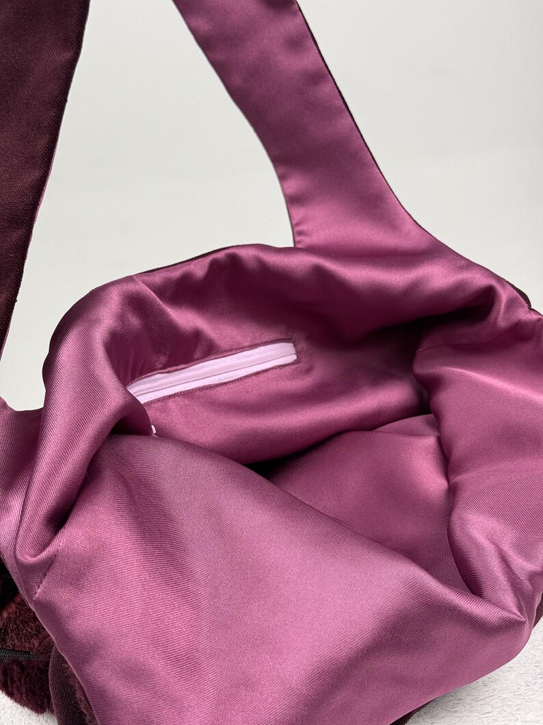 May include: A close-up of a tote bag with a deep burgundy exterior and a satin-like, mauve interior. The bag has a long strap and a zipper closure. The fabric has a soft, luxurious appearance.