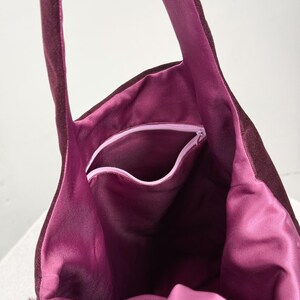 May include: A burgundy tote bag with a plush, velvety exterior and a vibrant magenta interior. The bag features a zippered pocket and long straps. The bag's interior is visible, showcasing its spacious design and contrasting color.