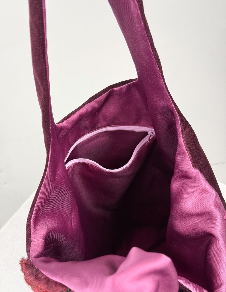 May include: A burgundy tote bag with a plush, velvety exterior and a vibrant magenta interior. The bag features a zippered pocket and long straps. The bag's interior is visible, showcasing its spacious design and contrasting color.
