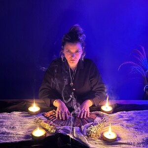 May include: A person sits at a table with tarot cards, surrounded by lit candles and incense. The scene is bathed in a blue light, with smoke rising from the incense and the person's hands. The person is wearing a dark shirt and jewelry.