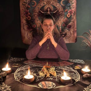May include: A person in a purple sweater with hands clasped in prayer, surrounded by candles and mystical items. A tapestry with floral and celestial designs hangs in the background. The scene evokes a sense of spirituality and divination.