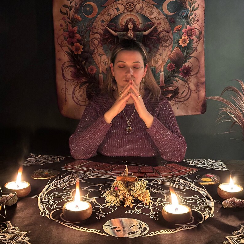 May include: A person in a purple sweater with hands clasped in prayer, surrounded by candles and mystical items. A tapestry with floral and celestial designs hangs in the background. The scene evokes a sense of spirituality and divination.