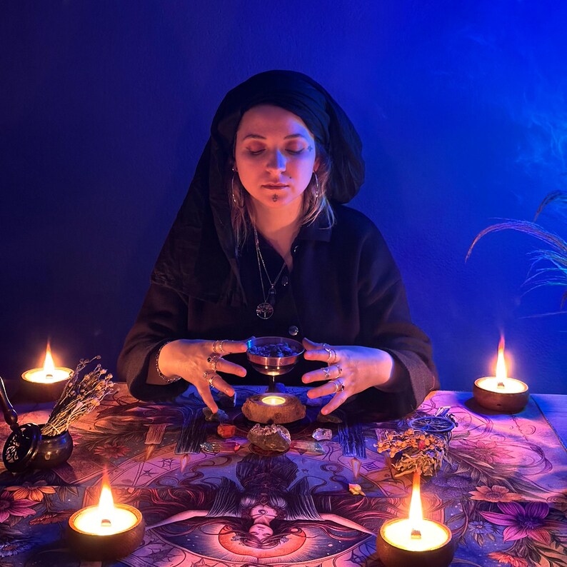 May include: A person in a black head covering and dark clothing sits at a table with lit candles and a patterned cloth. A small bowl is held above a candle, with other items on the table, creating a mystical atmosphere.